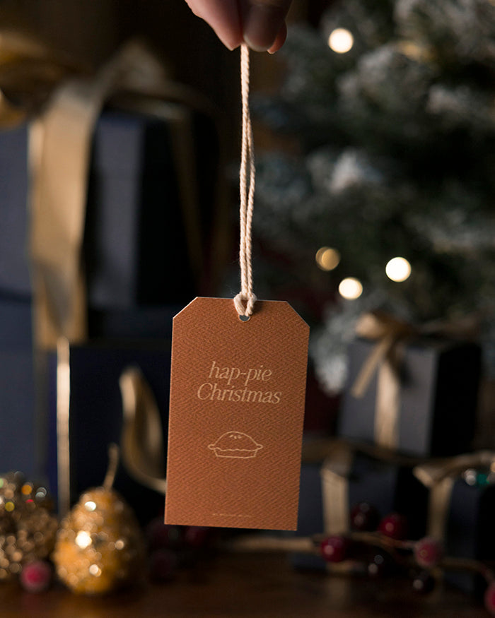 My Brunch Plans' gift tag with 'hap-pie Christmas' text and pie illustration, held by a hand against a festive background.