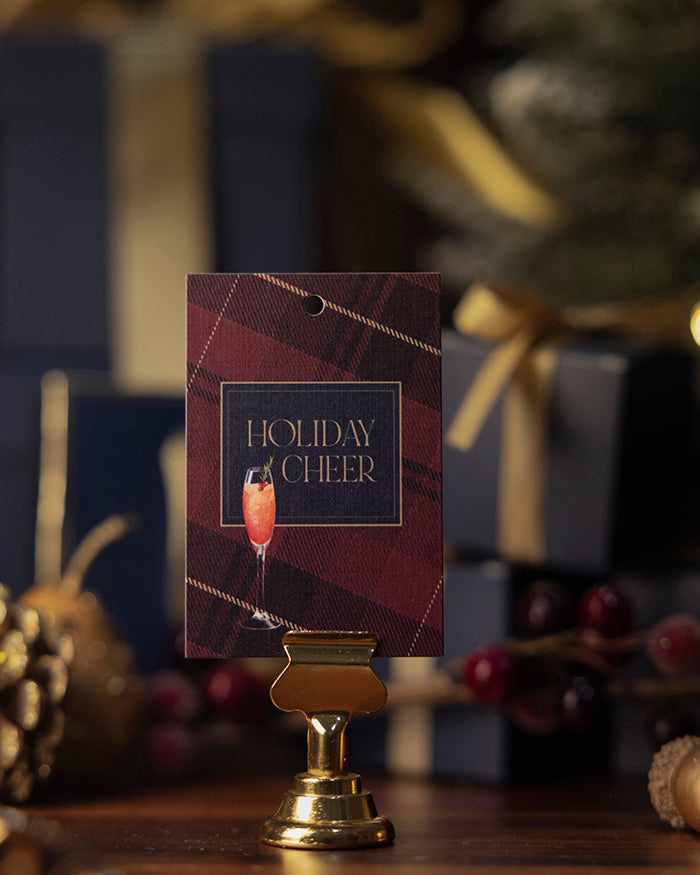My Brunch Plans' tartan linen gift tag with 'Holiday Cheer' text and a cocktail illustration, placed on a gold stand with a festive background.