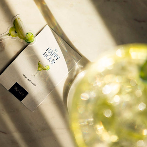 a flatlay of My Brunch Plans’ hand painted gouache martini diecut sticker along with a sweet notes affirmation card reading " it's happy hour when you decide it is" on a marble table with a martini glass in the afternoon golden sunlight