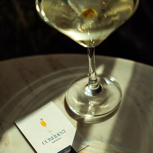 a dirty martini cocktail glass on a marble table with a My Brunch Plans' sweet note affirmation card that reads "be confident bottomless" and a gouache painted mimosa.