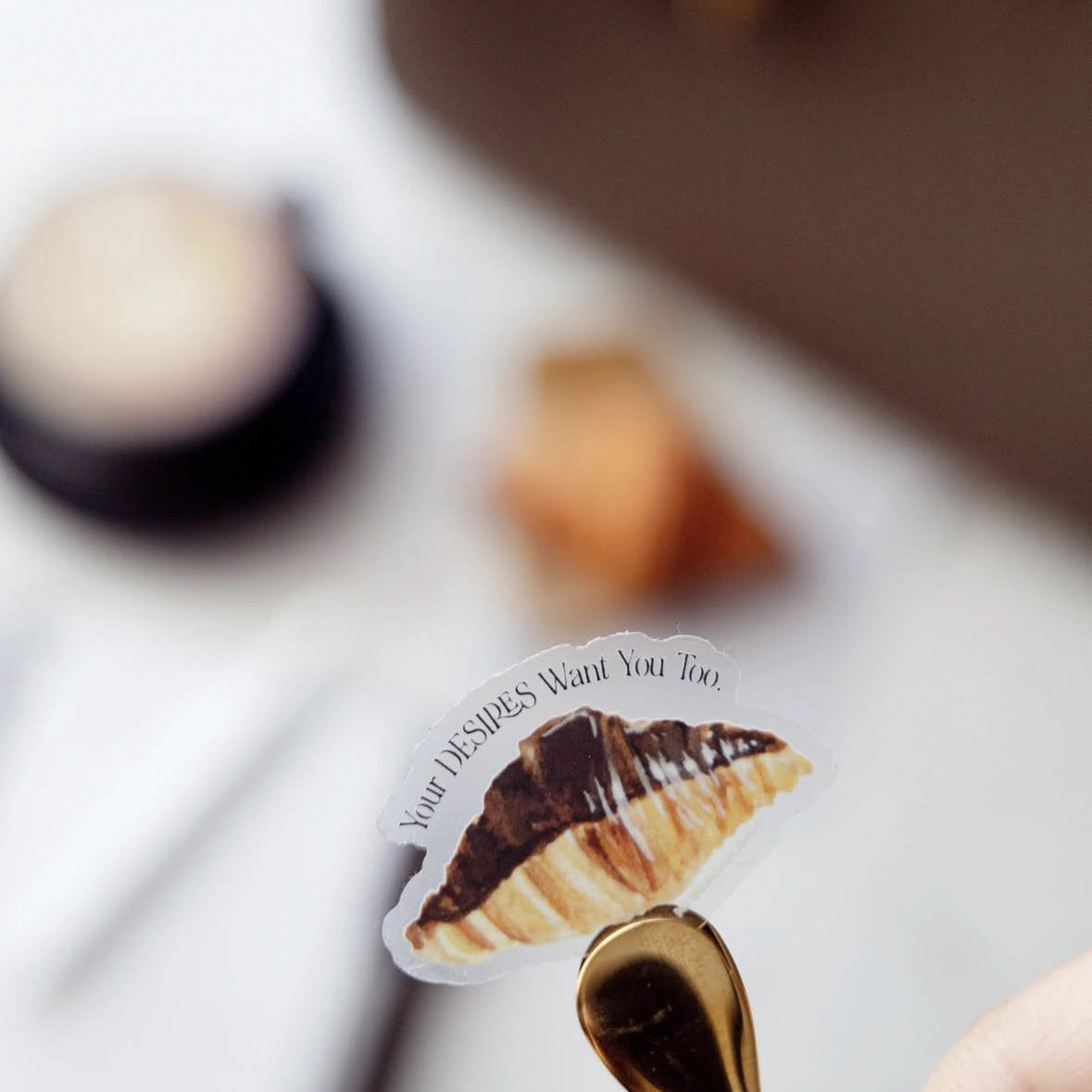 My Brunch Plans’ hand painted gouache chocolate covered croissant with the phrase "your desires want you too" diecut sticker with a latte, croissant and handbag in the background.
