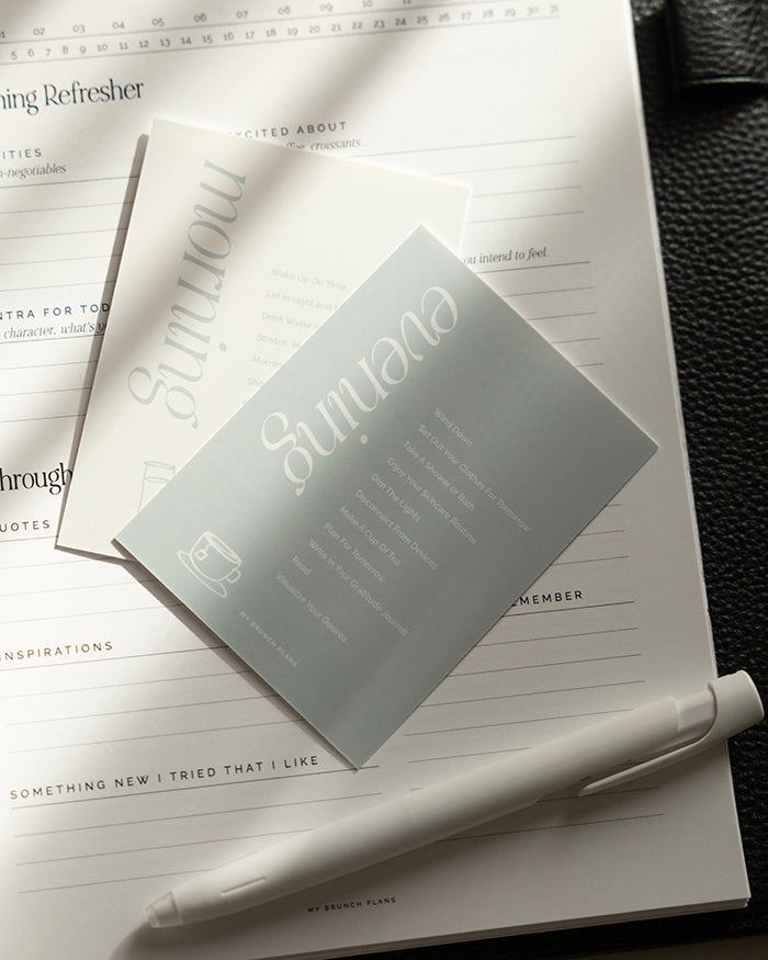 Close up of My Brunch Plans' planner card set of two. One is a morning routine suggestion with a hand drawn line art of lemon water with a white background and blue text. The evening routine card has hand drawn line art of a cup of tea with the same blue but as the background. Cards are set on a planner with a pen.