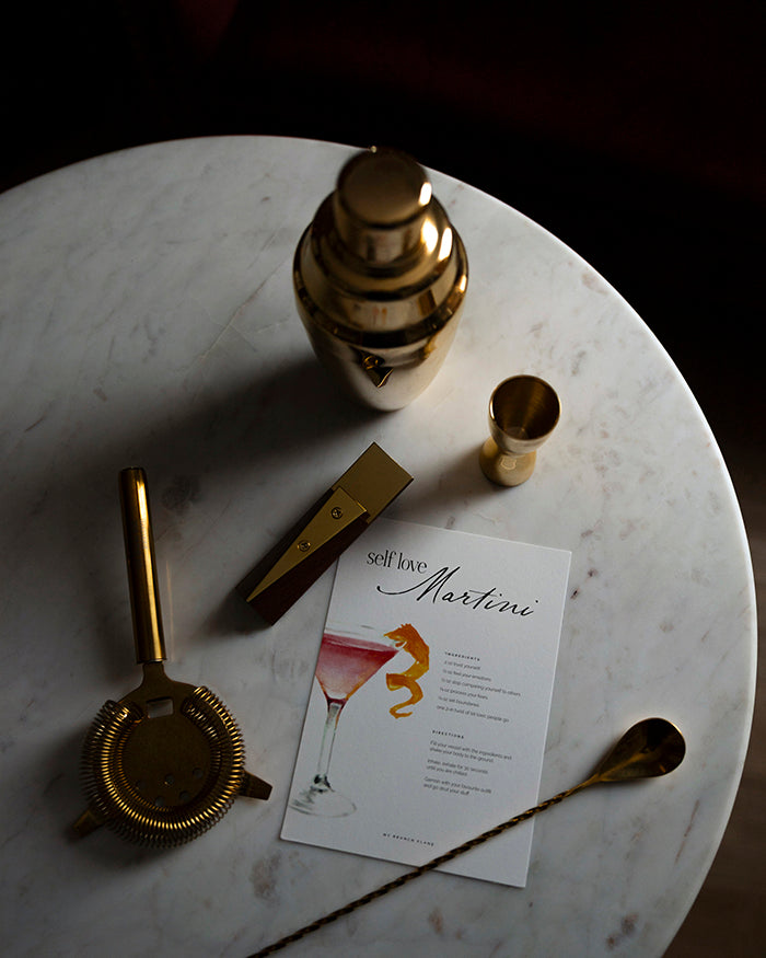 Gold cocktail shaker, strainer, and jigger on a marble surface with a cocktail recipe card.