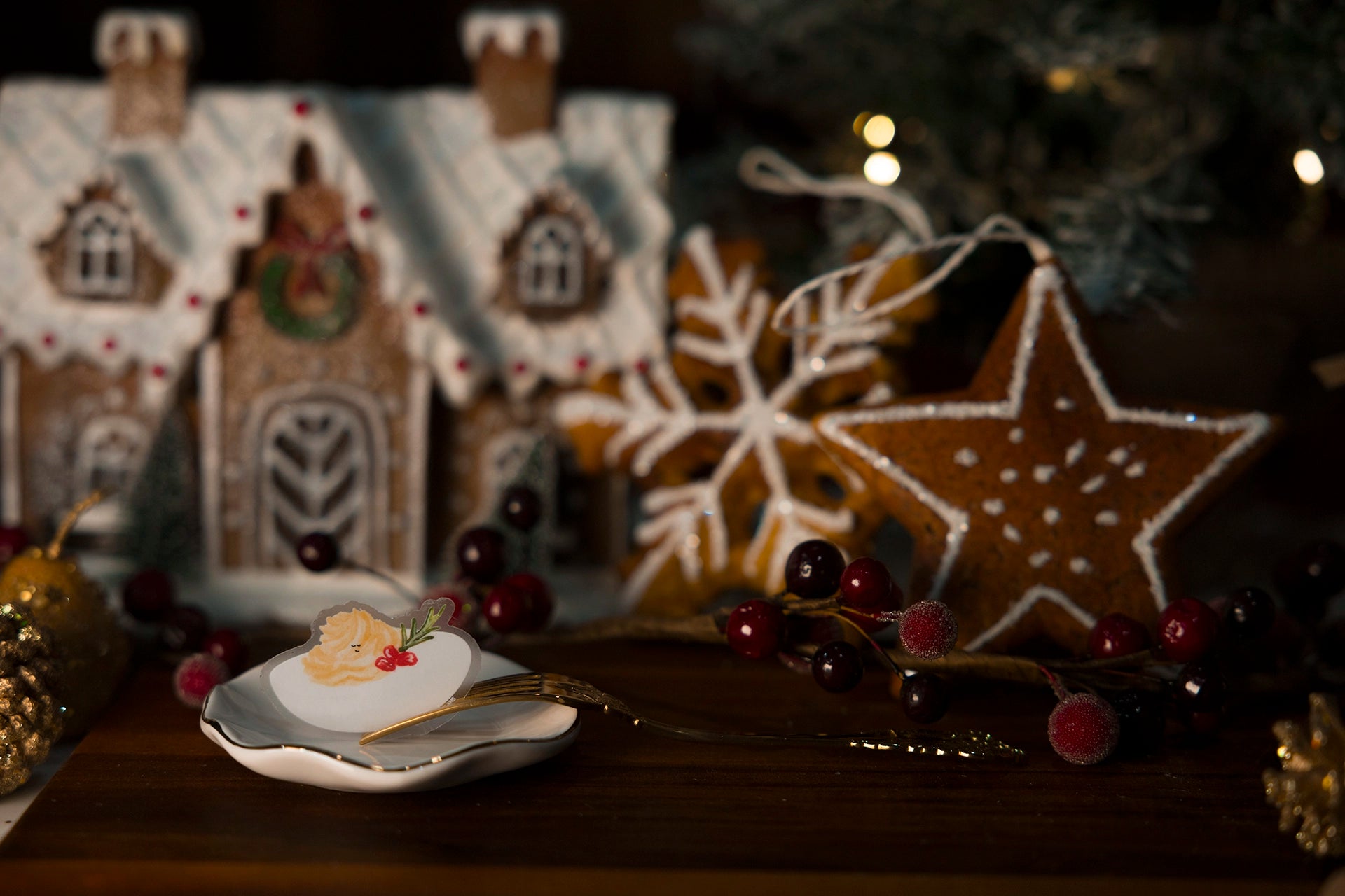 Decorative gingerbread houses and star with berries on a wooden surface, Christmas tree in the background.