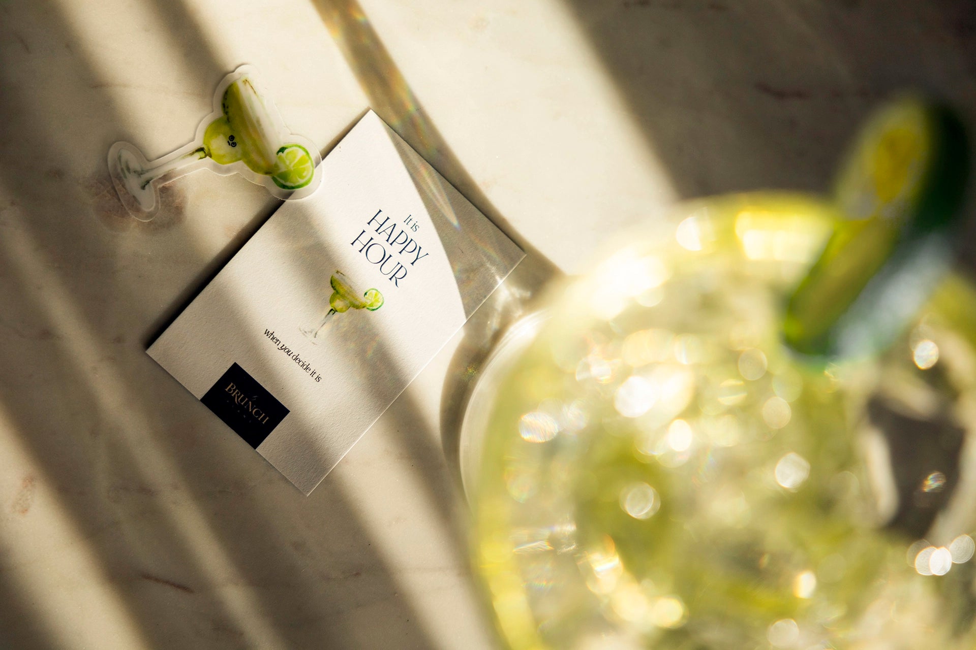 a flatlay of My Brunch Plans’ hand painted gouache  martini diecut sticker along with a sweet notes affirmation card reading " it's happy hour when you decide it is" on a marble table with a martini glass in the afternoon golden sunlight