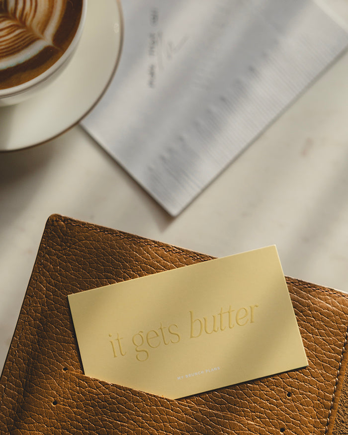 My Brunch Plans' planner card with the phrase "it get's butter" in a raised gloss tone on tone on a butter yellow card tucked in a brown planner card slot with a cup of latte and our napkin notepad in the background set on a marble table.