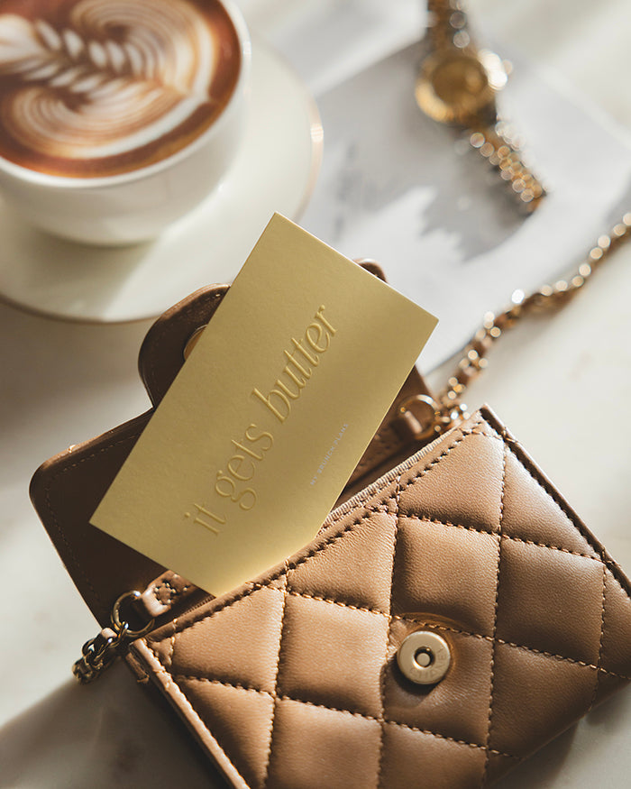My Brunch Plans' planner card with the phrase "it get's butter" in a raised gloss tone on tone on a butter yellow card tucked in a luxurious brown leather quilted wallet on chain with a cup of latte, a luxury watch and our napkin notepad in the background set on a marble table.