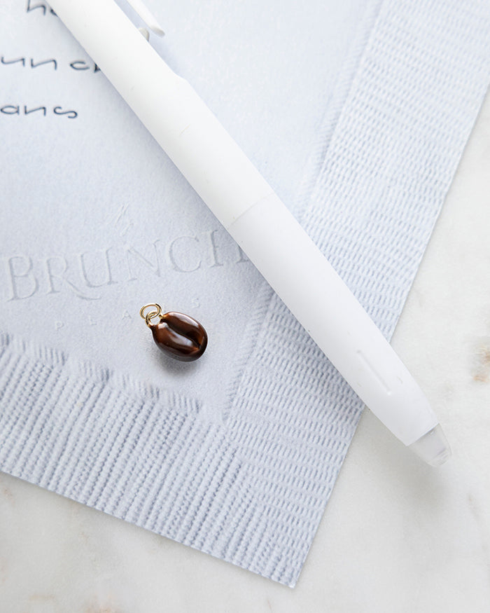 Coffee Bean Charm | 18k Gold Plated with Enamel on My Brunch Plans’ Napkin notepad with a pen for size comparison.